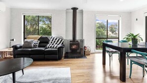 A modern living room with a black leather sofa, a dining table with chairs, a fireplace, and large windows offering a view of greenery. - Orana - Free Wi-Fi 2 blocks to Beach (Apollo Bay)