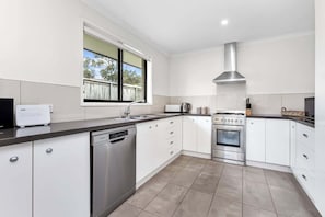 A modern kitchen with white cabinetry, a dishwasher, an oven, and a window with a view of the outdoors.