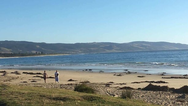 Uma praia com duas pessoas caminhando, céu claro e colinas ao longe.