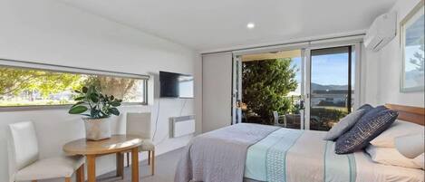 A modern bedroom with a bed, a small table, a chair, a potted plant, and a sliding glass door leading to a balcony.