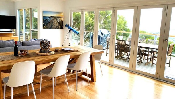 A modern dining area with a wooden table and chairs, a telescope, and a large window with a view of the outdoors. - Apollo Ocean Blue Ocean View Wi-Fi Pet Friendly (Apollo Bay)