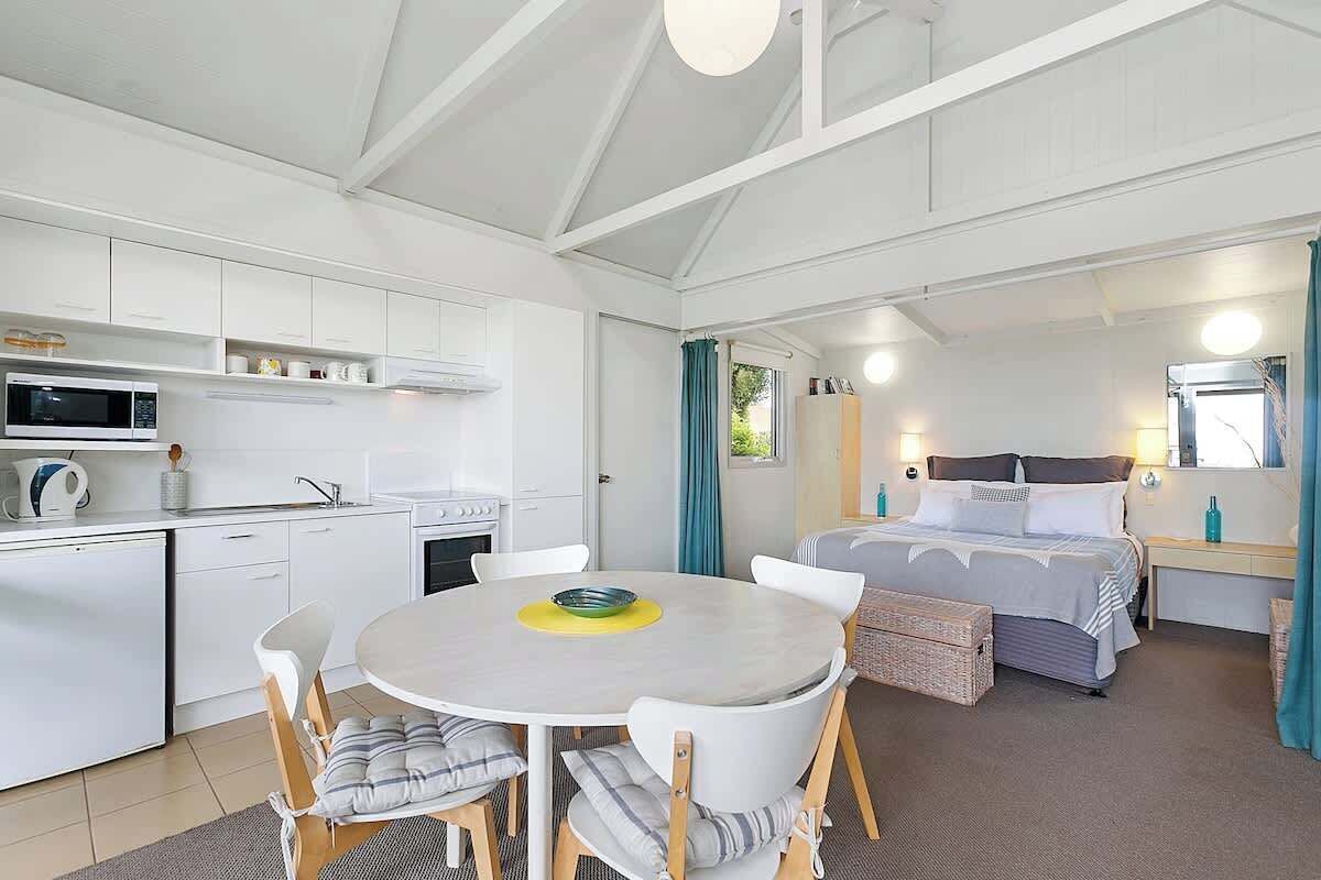 Une pièce compacte comprenant un coin cuisine, une table de salle à manger ronde avec des chaises, un lit avec une tête de lit et une fenêtre avec des rideaux.