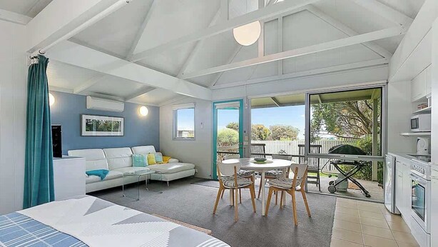 Una habitación moderna con cama, sofá, mesa de comedor con sillas, zona de cocina y ventanas amplias.