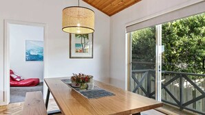 Une table à manger en bois, surmontée d’un luminaire suspendu en fibres tressées, avec un tableau encadré et une vue sur un balcon doté d’une balustrade.