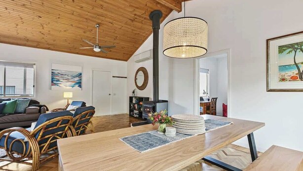 Un vaste espace de vie doté d’un plafond en bois, d’une table à manger agrémentée d’un centre de table en fibres naturelles, de meubles en osier et d’un grand tableau mural.