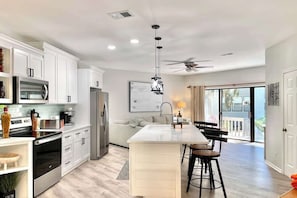 Une cuisine moderne avec des meubles blancs, des appareils en acier inoxydable, un îlot central doté de tabourets et un ventilateur de plafond.