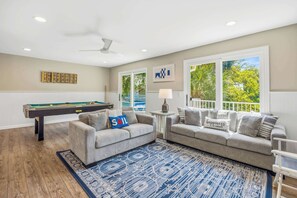 Uma sala de estar com mesa de bilhar, dois sofás, um ventilador de teto e vista para o exterior.