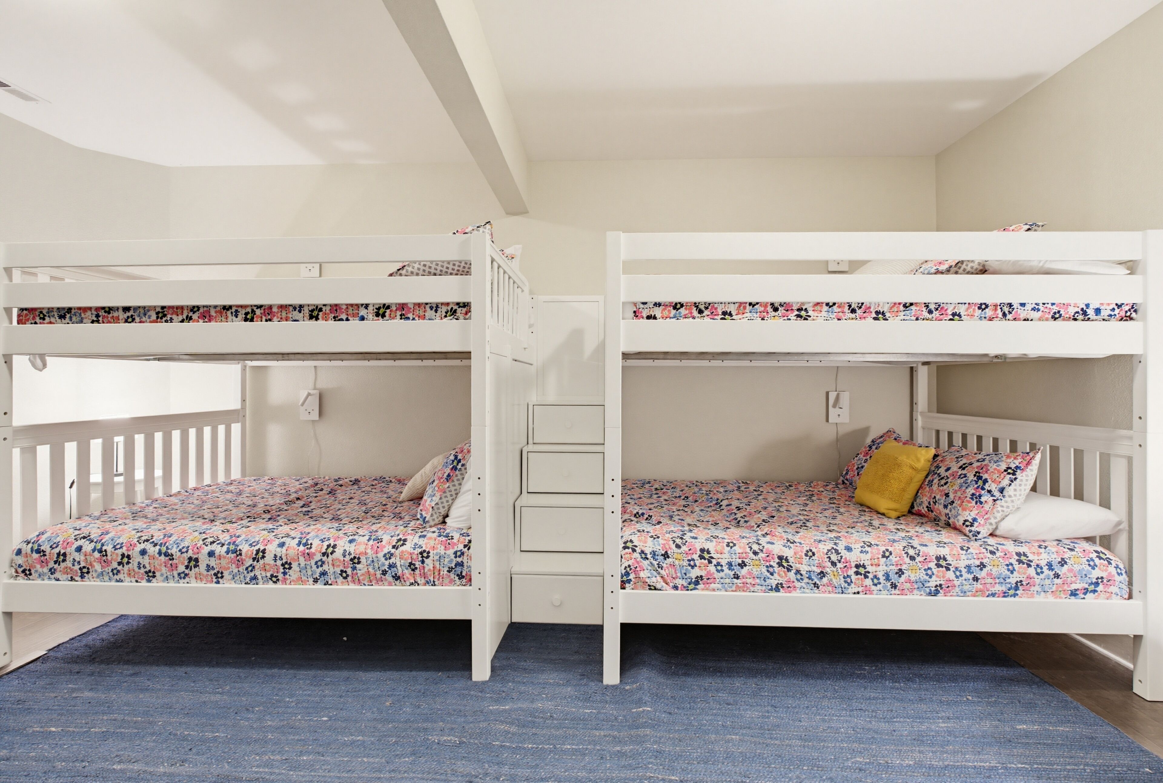 A room with two bunk beds, each with a floral-patterned mattress and a white headboard. There is a blue carpet on the floor, and a white wall with a power outlet.