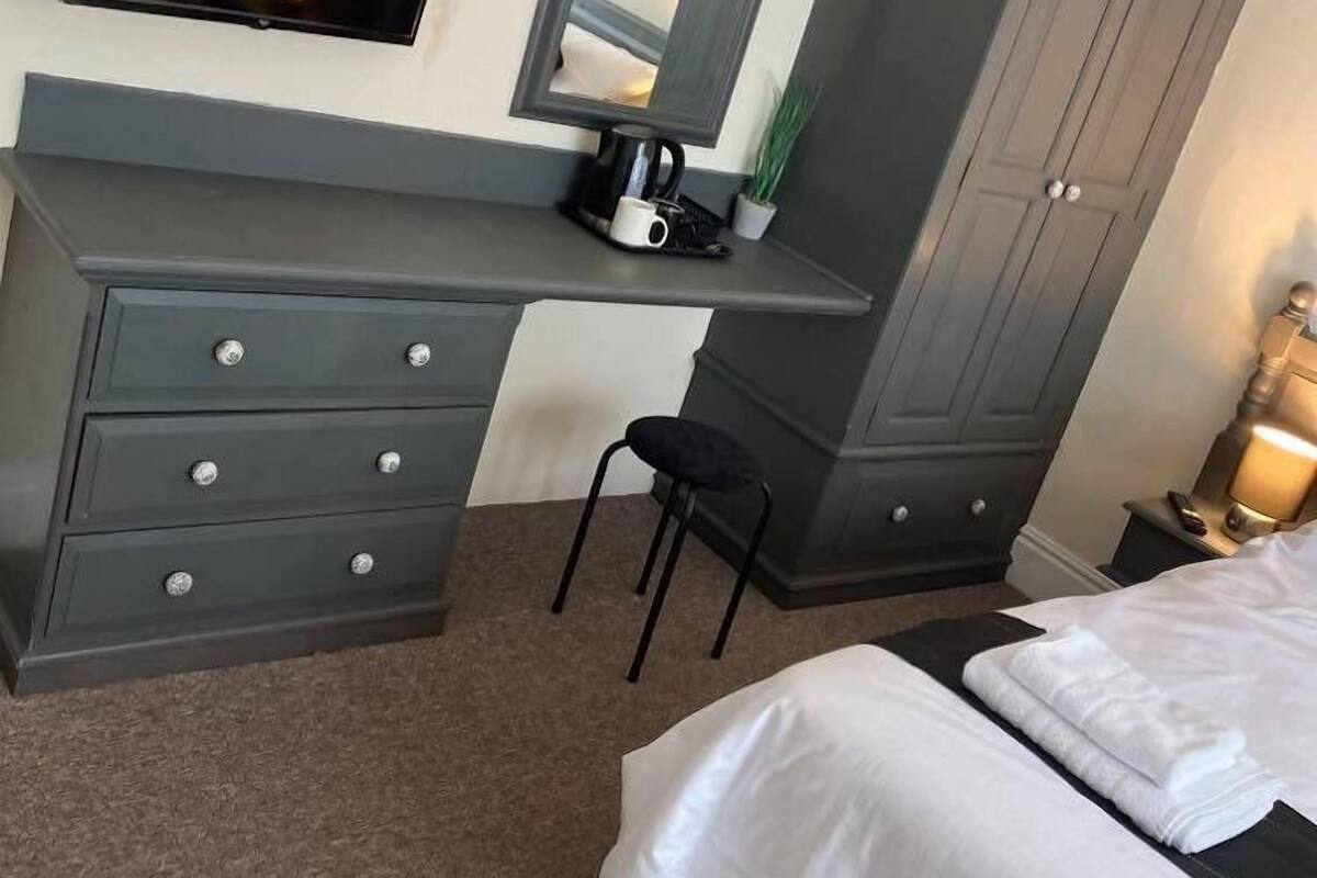A bedroom with a bed, a desk with drawers, a chair, a wardrobe, a TV, and a bedside lamp.