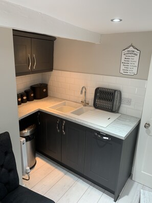 A compact kitchen with dark cabinets, a white countertop, a sink, and a stainless steel trash bin. A sign on the wall indicates the kitchen's opening hours.