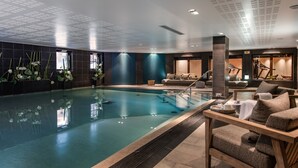 Zona d'una piscina interior moderna amb una cadira de relaxació de fusta, una taula petita amb una tovallola i una paret decorada amb flors.