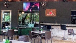 Uma área de refeições ao ar livre com mesas, cadeiras, um balcão de bar e uma tela de TV exibindo um programa.