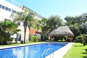 Una piscina con una estructura de techo de paja, rodeada de palmeras y un edificio con fachada blanca.