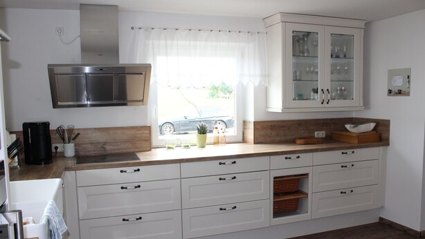 Una cocina moderna con armarios blancos, una campana extractora de acero inoxidable, una ventana con cortinas translúcidas y vista a un coche estacionado afuera.