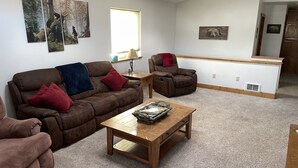 A living room with a brown leather couch, a wooden coffee table, and a red chair. There are wall-mounted pictures and a window with blinds. - Beartracks Retreat (Dubois)