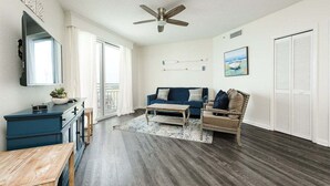 A spacious living room with a dark wood floor, a blue sofa, a wooden coffee table, a wooden bench, and a ceiling fan. - Ocean View Oasis - Ocean Walk (Daytona Beach)