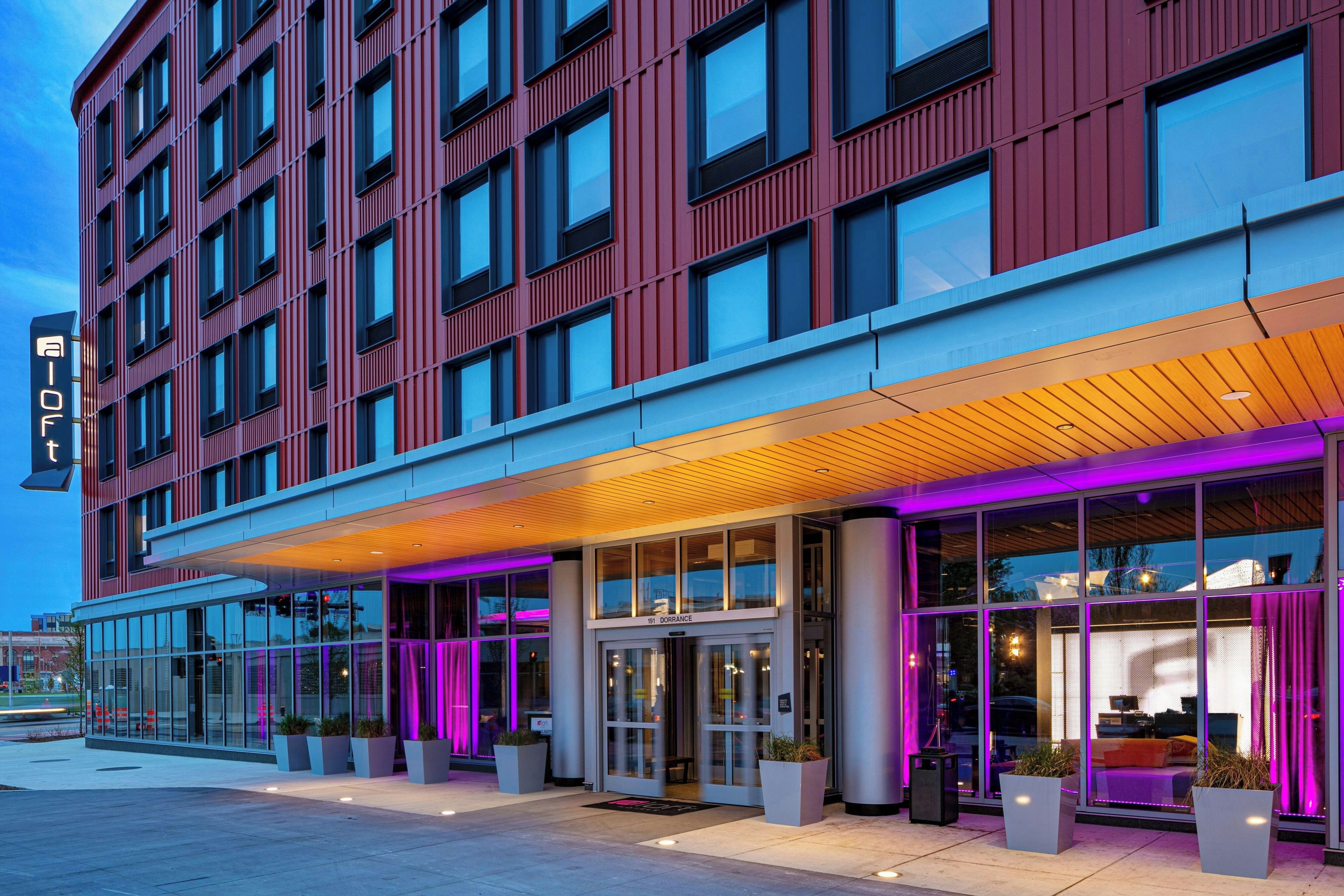 A modern hotel with a red facade, large windows, and a covered entrance area with purple lighting.