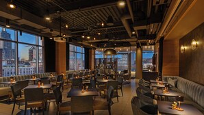 Un ristorante moderno con soffitti alti, condotti a vista e ampie finestre che offrono vista sulla città. Tavoli e sedie disposti per la ristorazione.