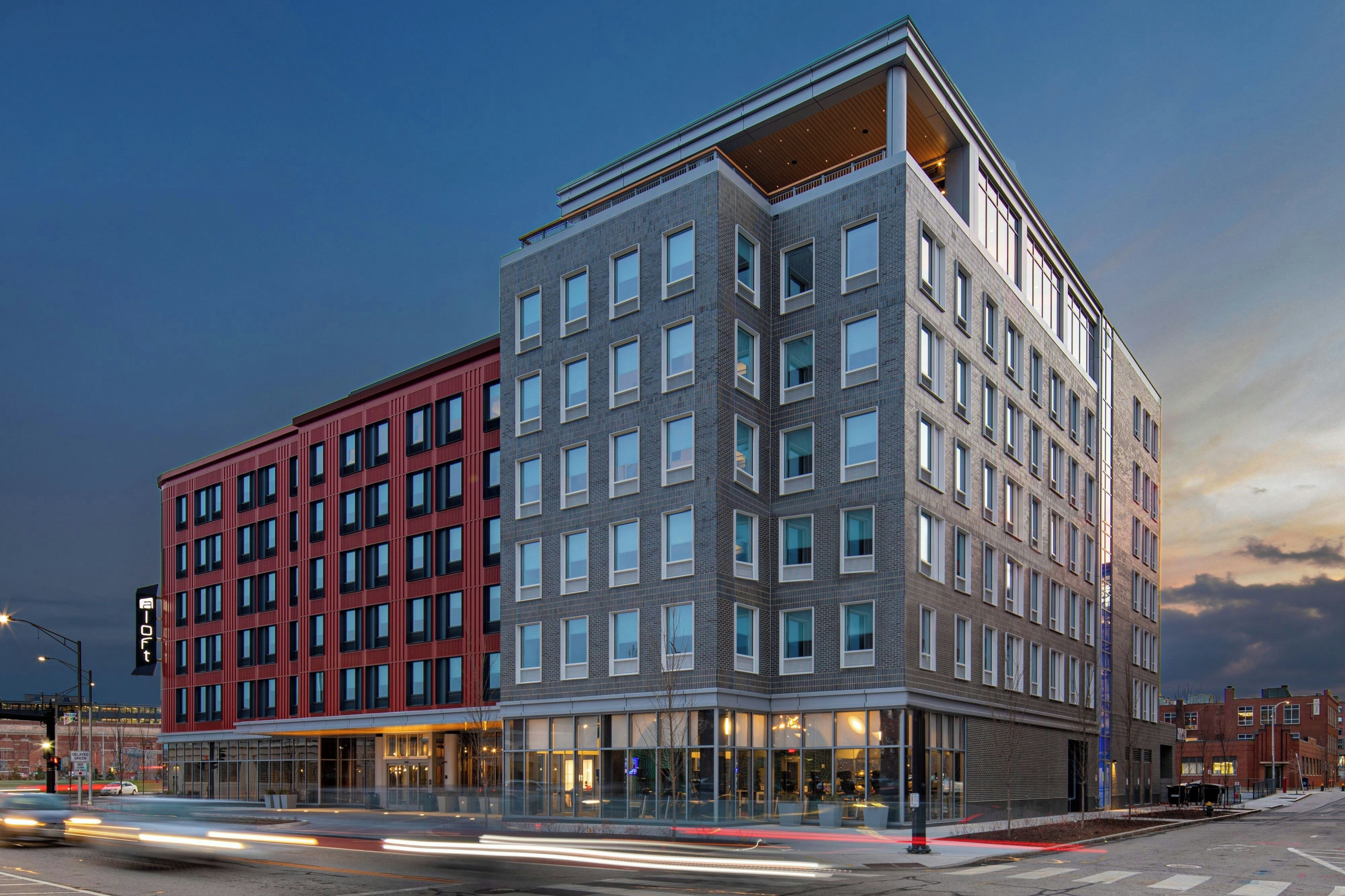 A multi-story hotel with a modern architectural design, featuring a mix of red and grey facades, large windows, and a corner entrance.