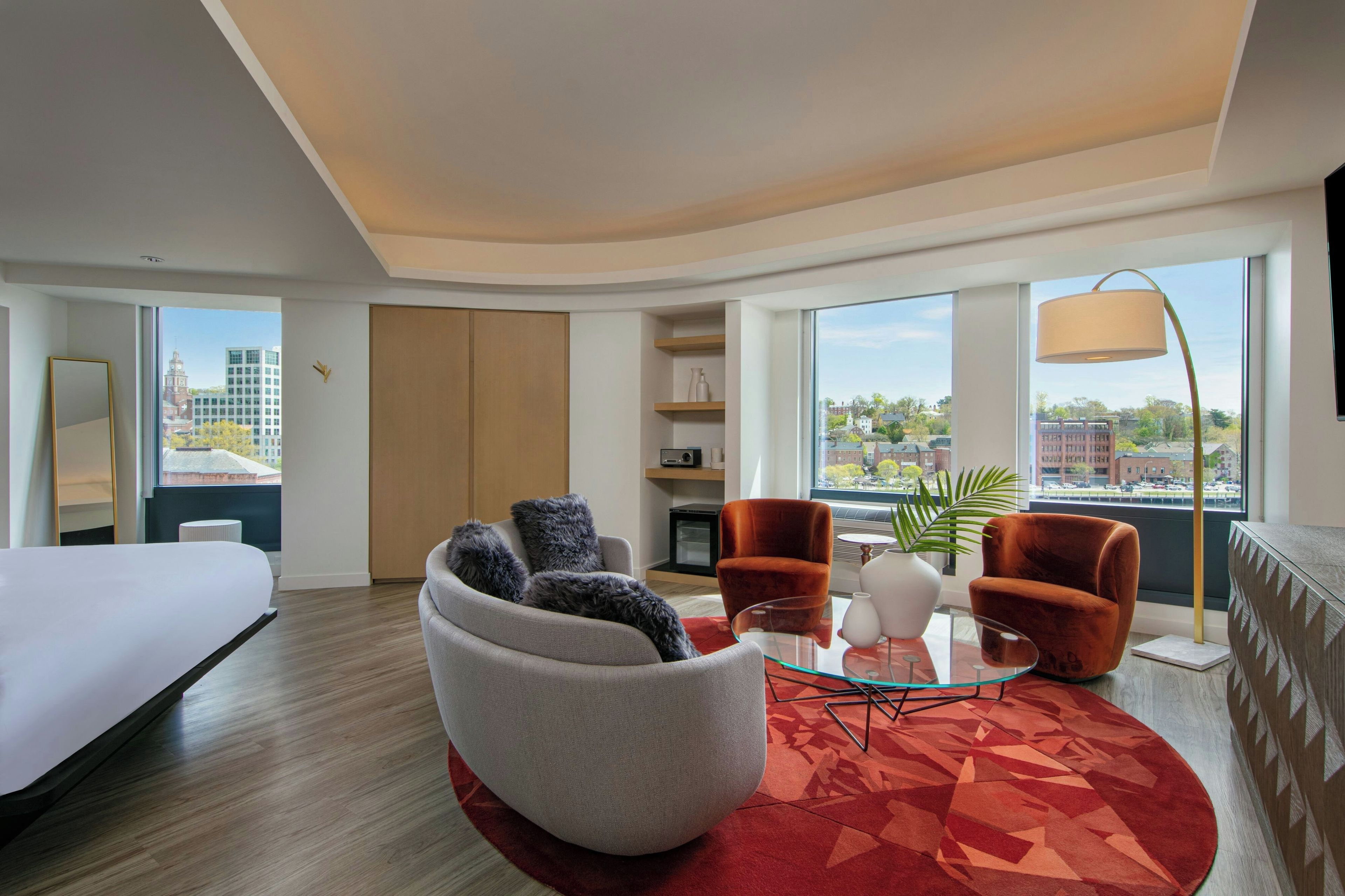 A modern hotel room with a bed, a seating area with a glass coffee table, and a view of the city.