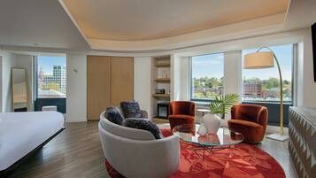 A modern hotel room with a bed, a seating area with a glass coffee table, and a view of the city.