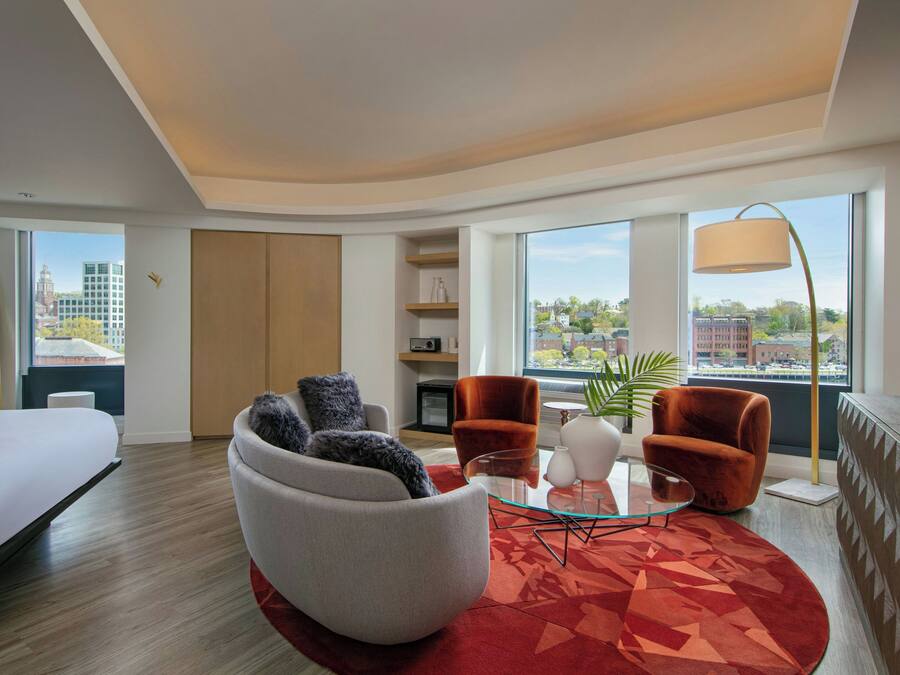 A modern hotel room with a bed, a seating area with a glass coffee table, and a view of the city.