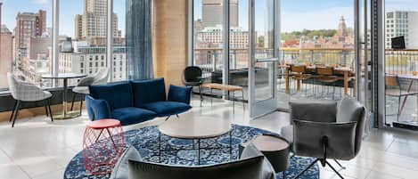 A modern lounge with a blue sofa, a round coffee table, and a red stool. There are multiple chairs and a dining area with a view of the cityscape.