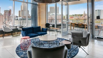 Un salón moderno con un sofá azul, una mesita de centro redonda y un taburete rojo. Hay varias sillas y un comedor con vistas a la ciudad.