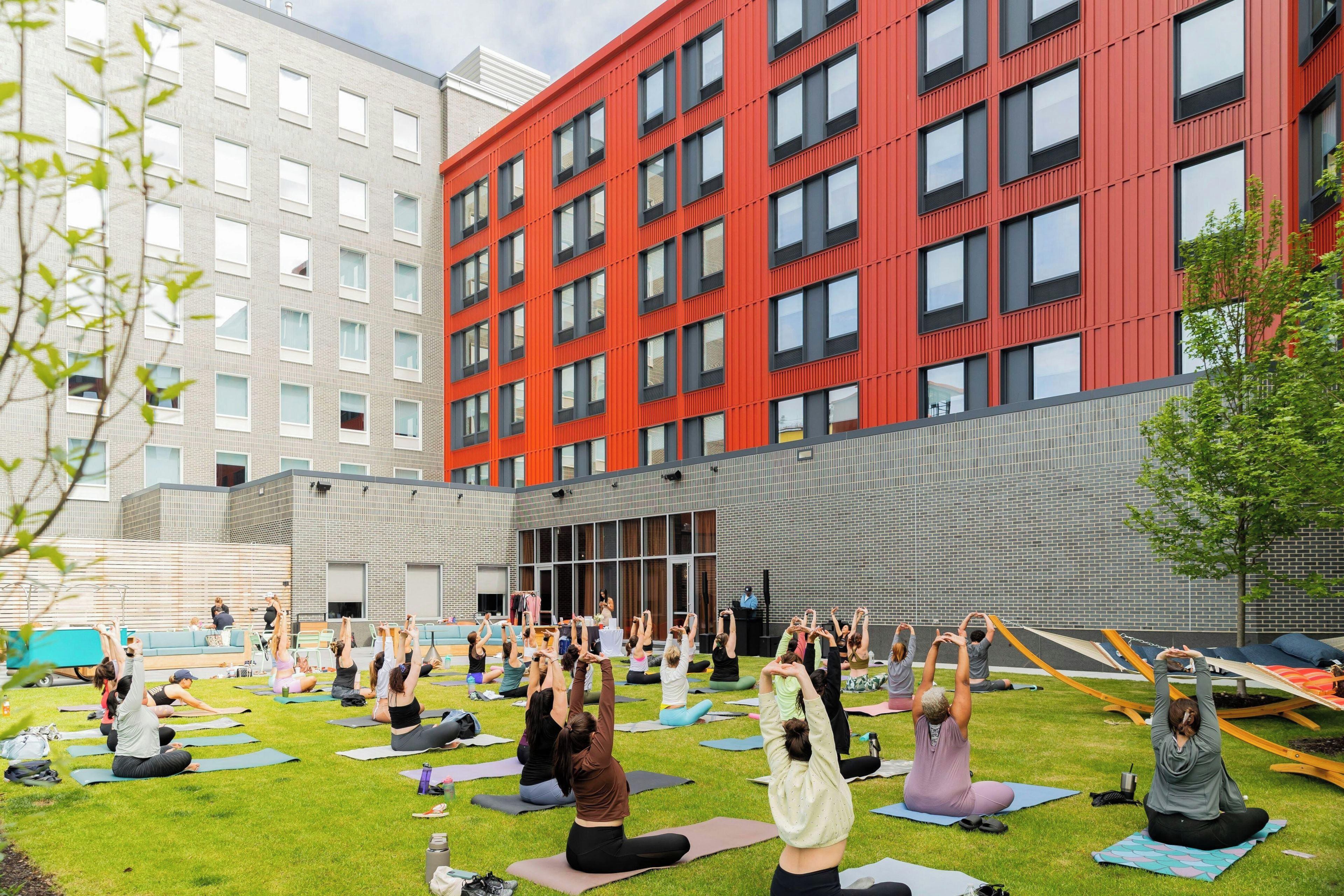 En grupp människor deltar i ett yogapass utomhus på en gräsyta, med en modern byggnad i bakgrunden.