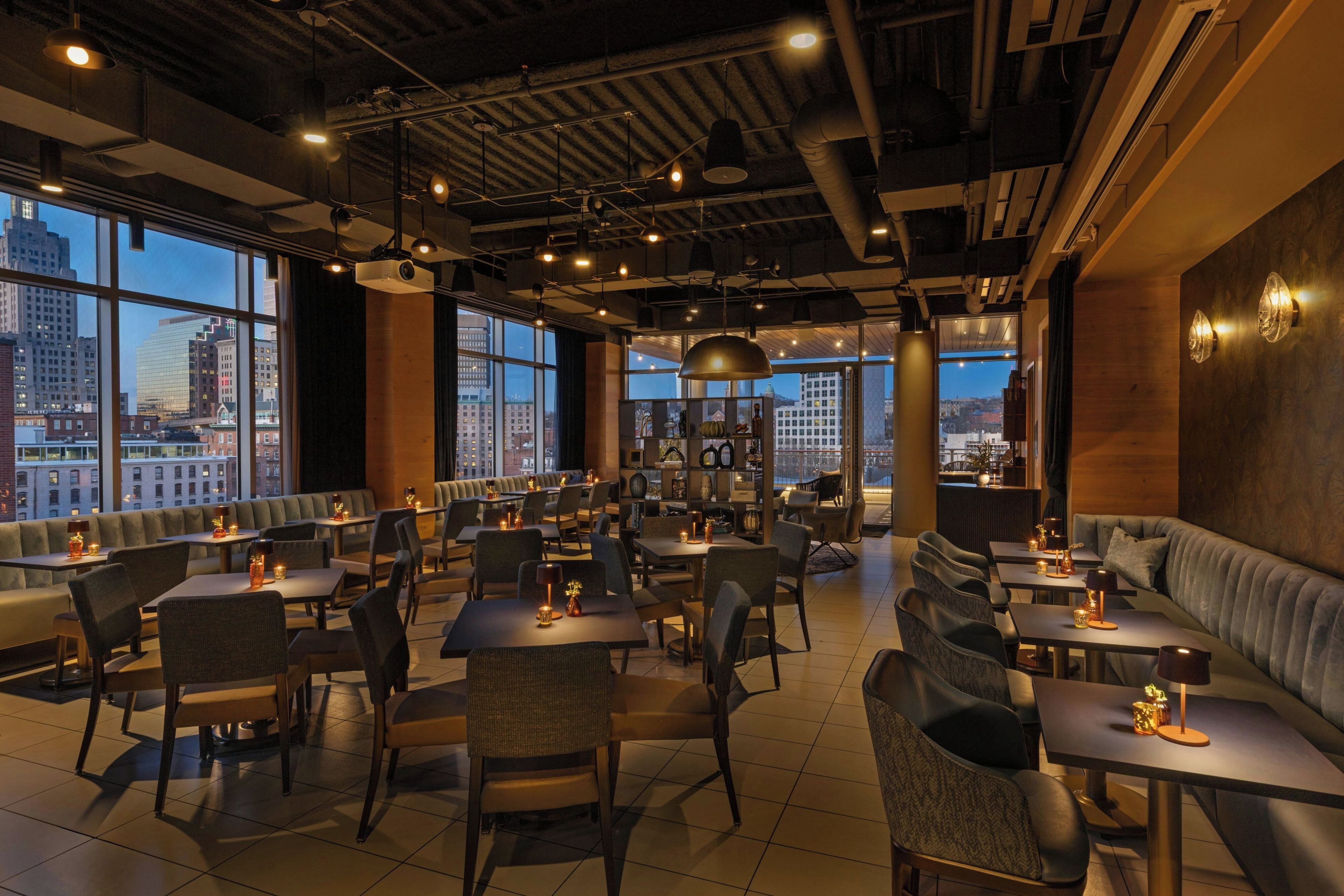 Een restaurant met hoge tafels en zitbanken, grote ramen met uitzicht op de stad, en een modern, industrieel plafond.