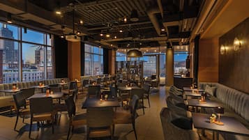 Een restaurant met hoge tafels en zitbanken, grote ramen met uitzicht op de stad, en een modern, industrieel plafond.