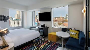 A modern hotel room with a large bed, a desk, a small table, and a sofa. There is a TV mounted on the wall and a window with a city view.