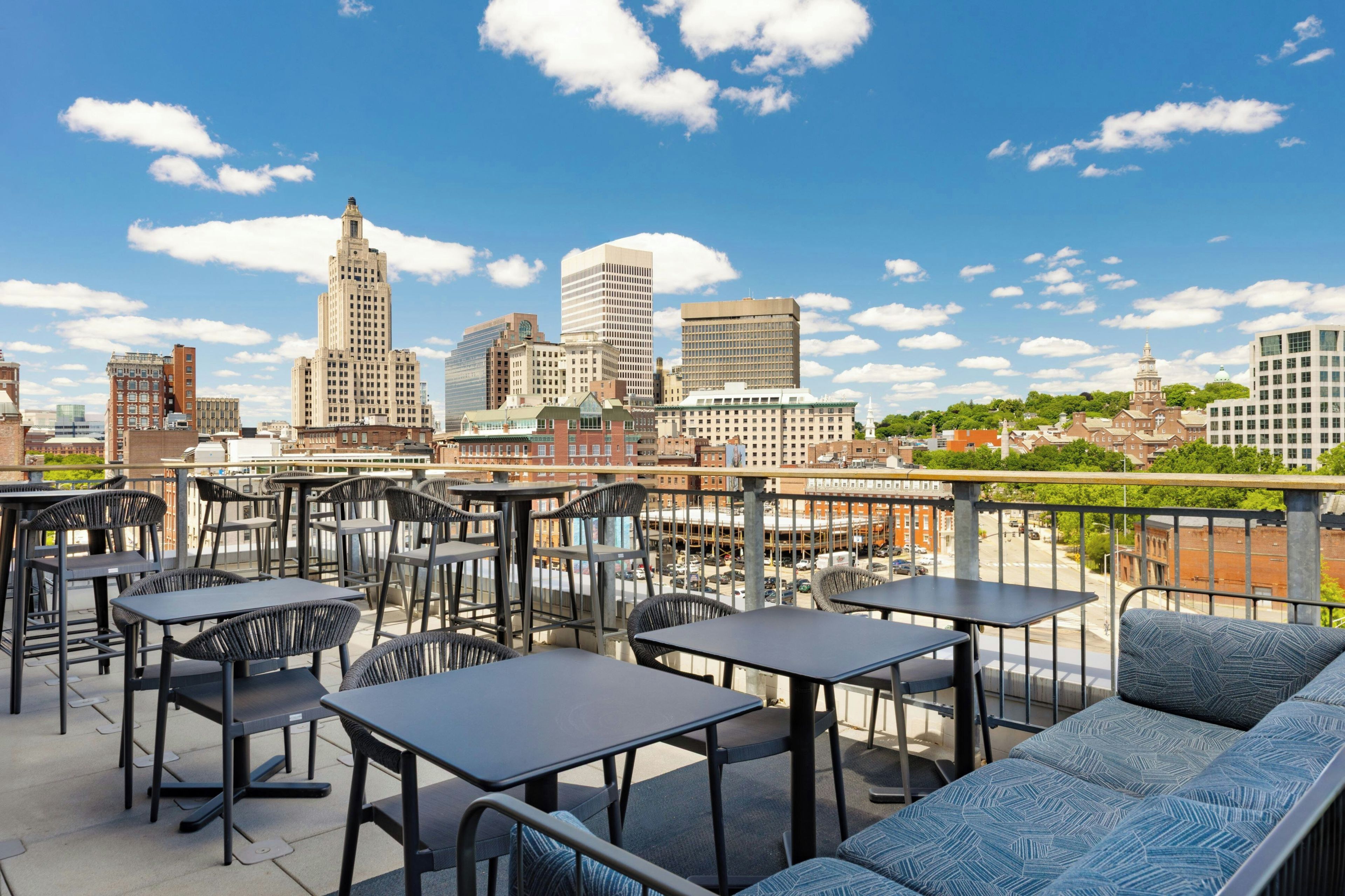A rooftop terrace with tables and chairs, overlooking a city skyline with various buildings.