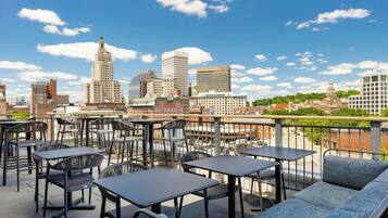 A rooftop terrace with tables and chairs, overlooking a city skyline with various buildings.