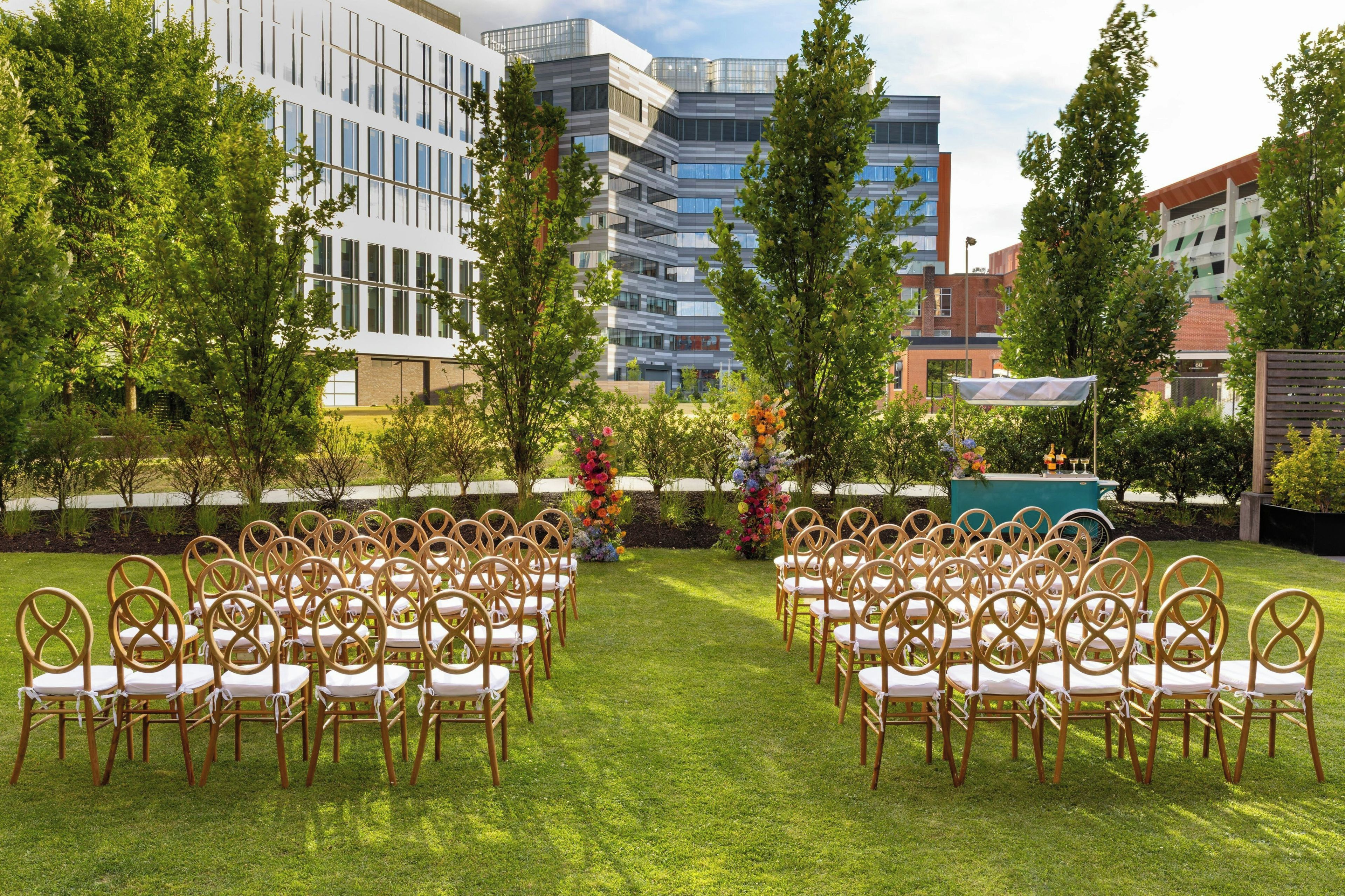 An outdoor seating area with rows of chairs and tables, surrounded by greenery and buildings in the background.