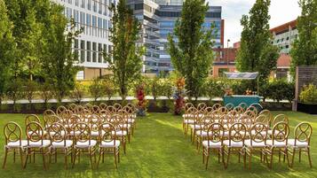 An outdoor seating area with rows of chairs and tables, surrounded by greenery and buildings in the background.