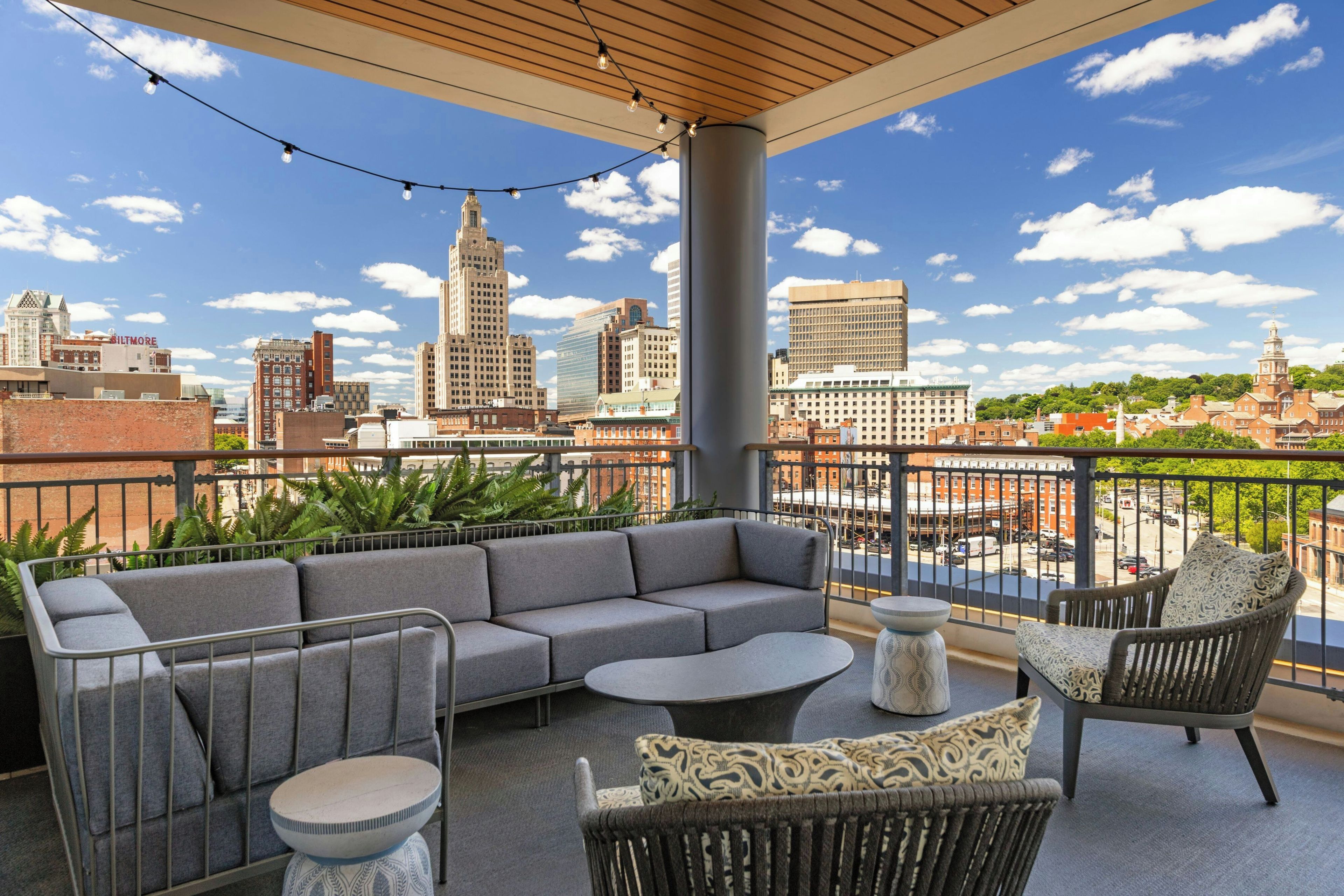 A balcony with a grey sofa, a round table, and wicker chairs, overlooking a cityscape with various buildings.