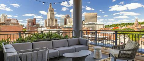 A balcony with a grey sofa, a round table, and wicker chairs, overlooking a cityscape with various buildings.