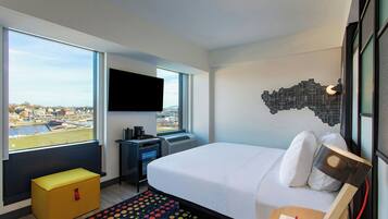 A hotel room with a large window, a bed, a desk, and a TV.