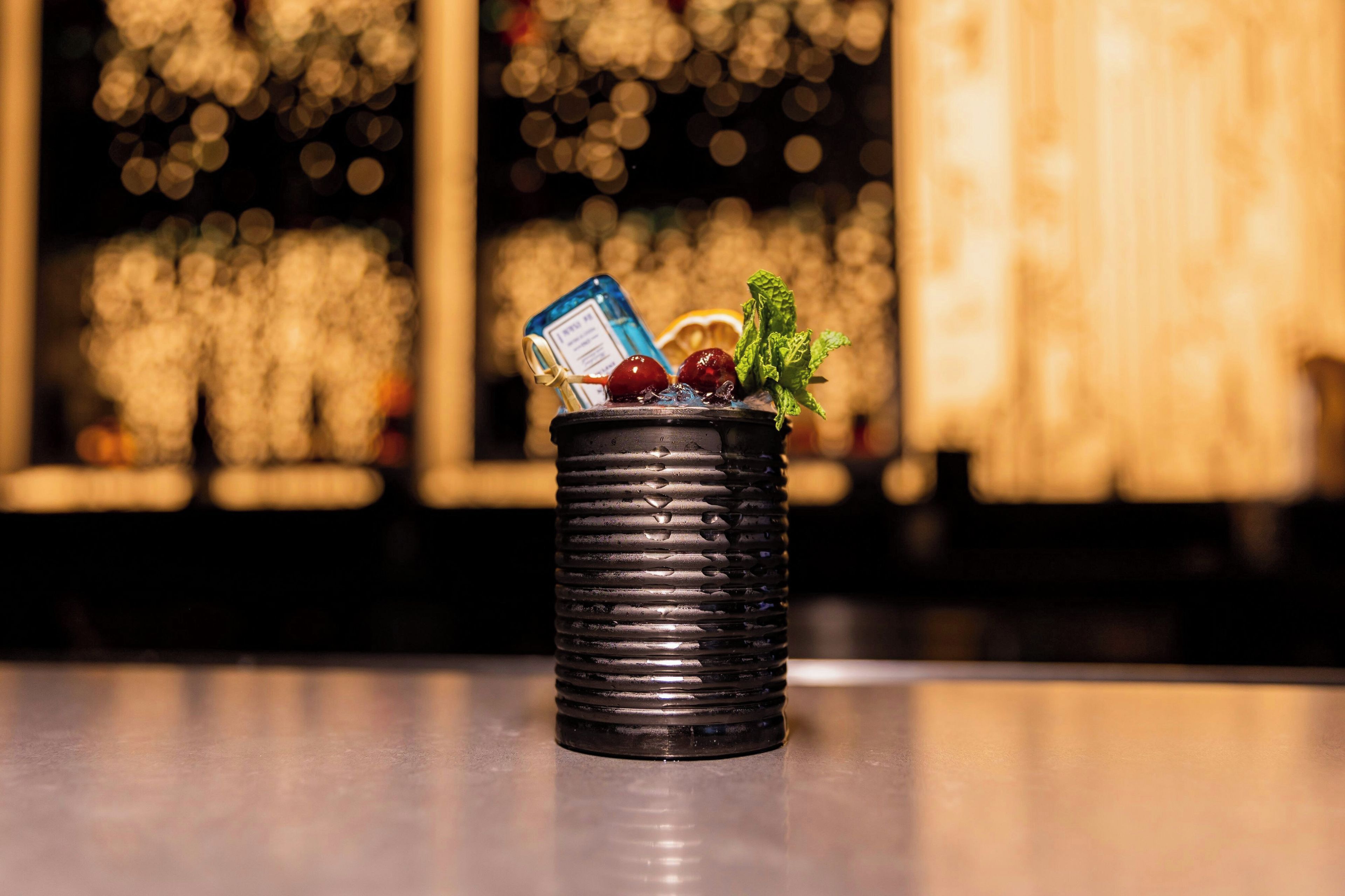 A black textured container with a blue bottle, a cherry, and mint leaves on a reflective surface.