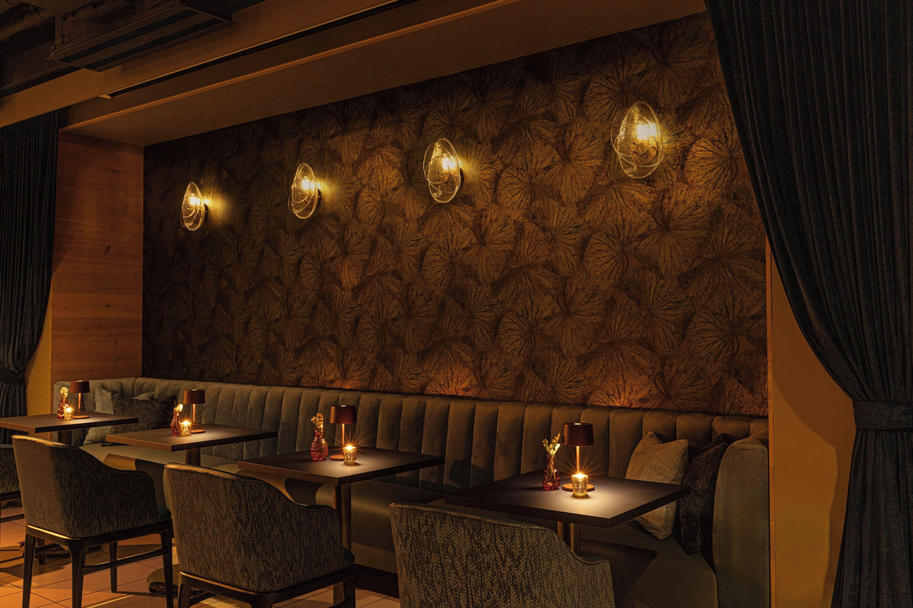 A dimly lit restaurant with a patterned wall, cushioned seating, and tables with candles.