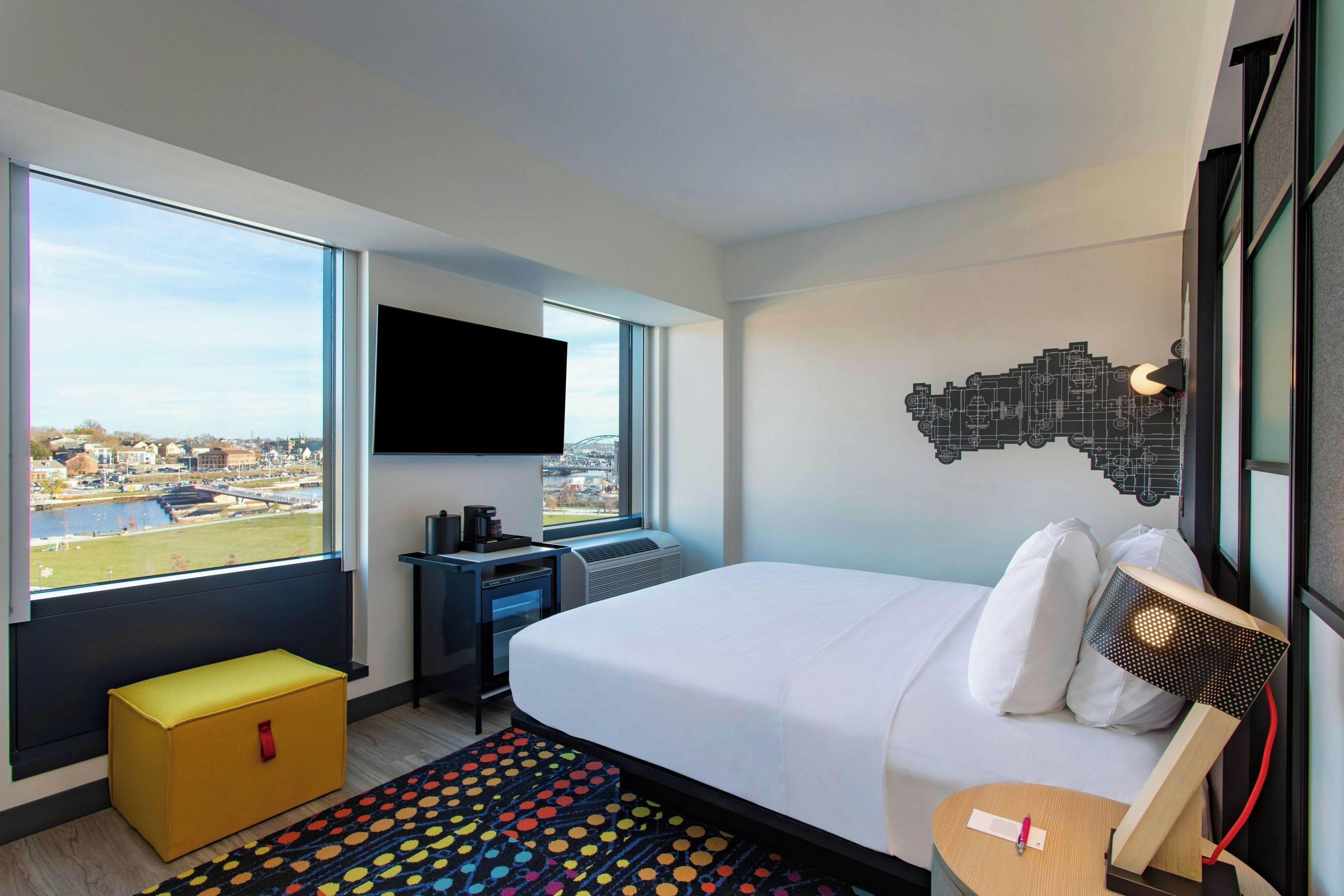 A hotel room with a bed, a desk, a TV, and a window with a city view.
