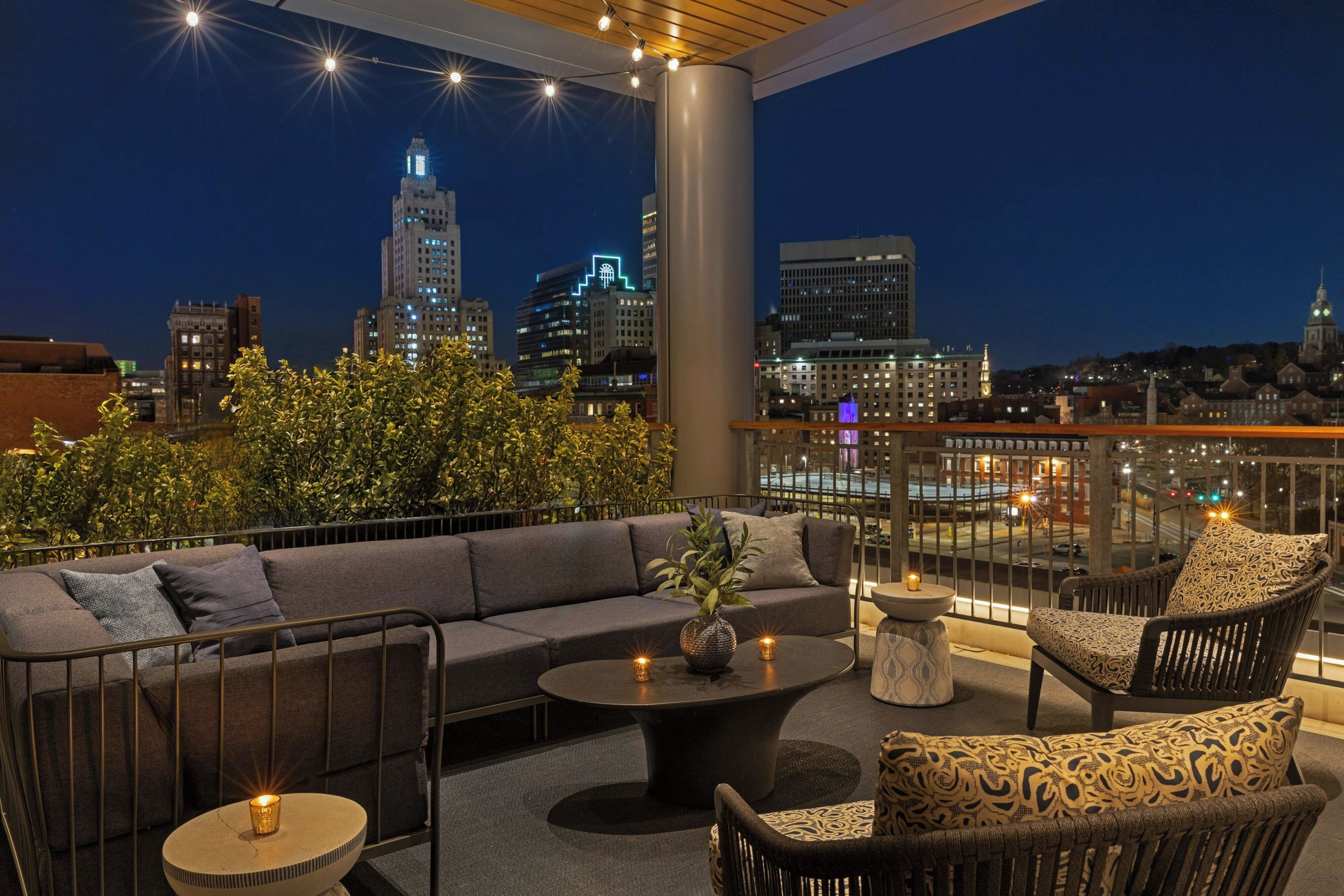 A rooftop terrace with a sofa, chairs, a coffee table, and a view of the city skyline at night.