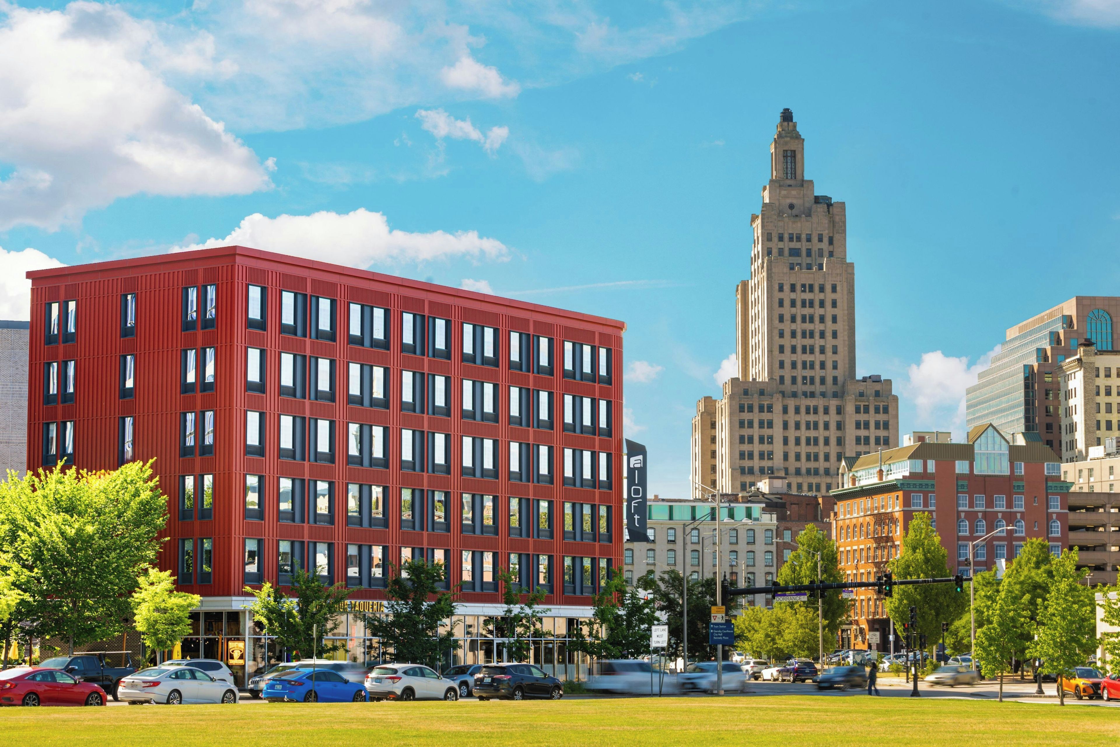 A modern red building with multiple windows, surrounded by a grassy area and other buildings.