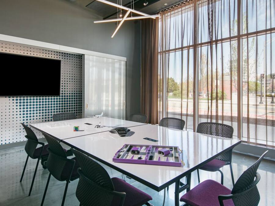 A modern conference room with a large glass table, black chairs, a mounted TV, and large windows with curtains.