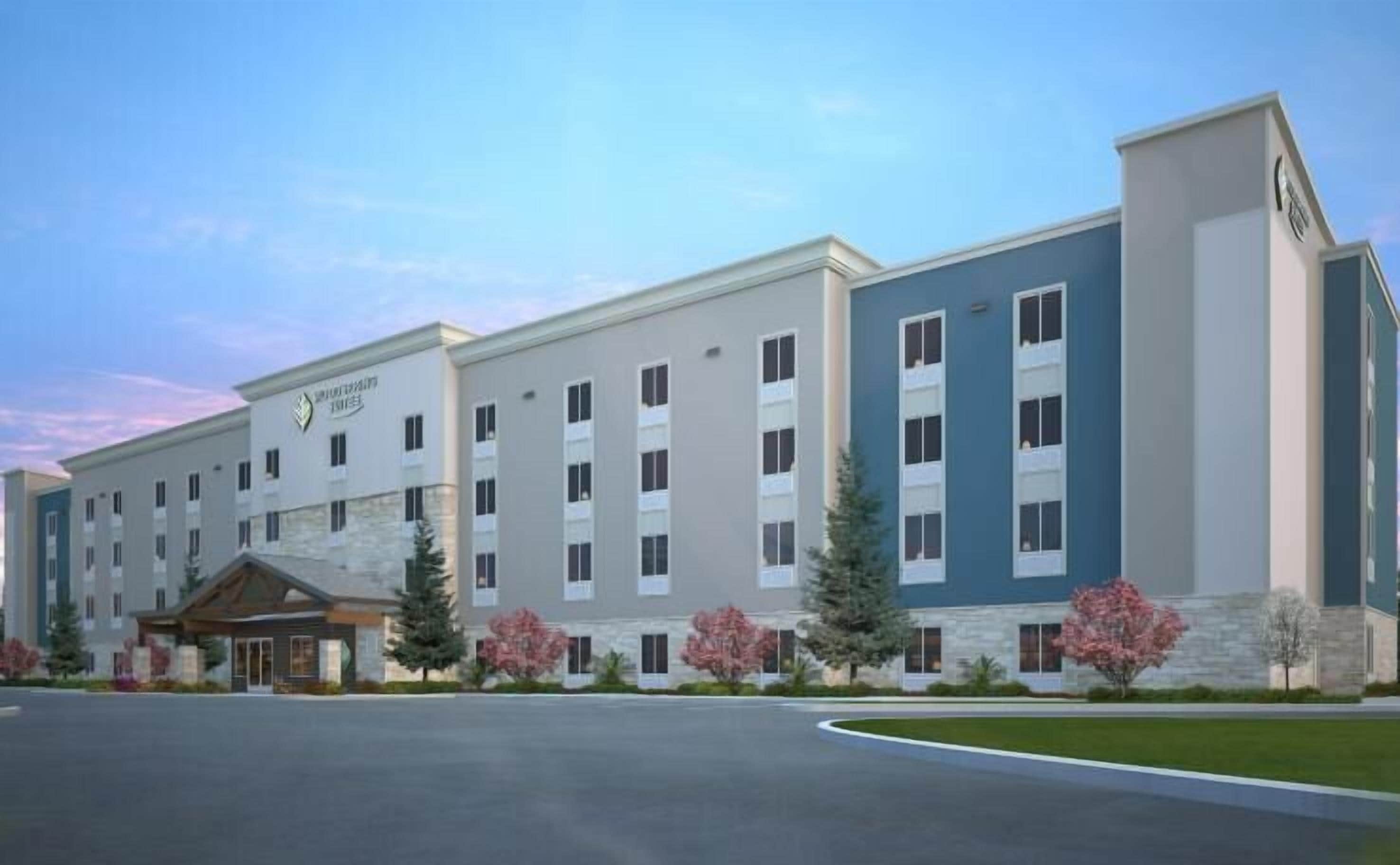 A multi-story hotel with a modern design, featuring a central entrance, multiple windows, and a landscaped front yard with trees and shrubs.