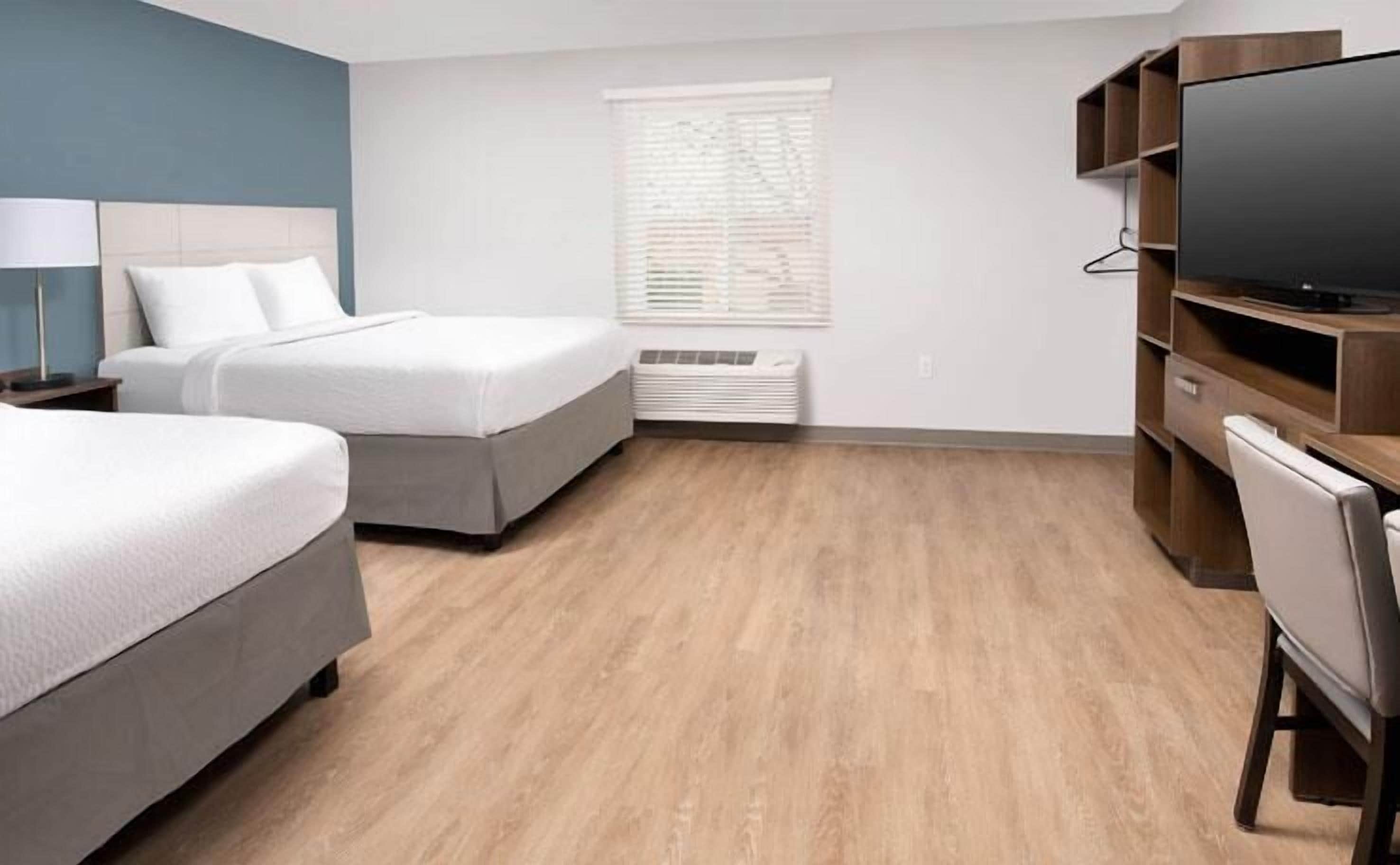 A hotel room with two beds, a TV, a desk, and a window with blinds.