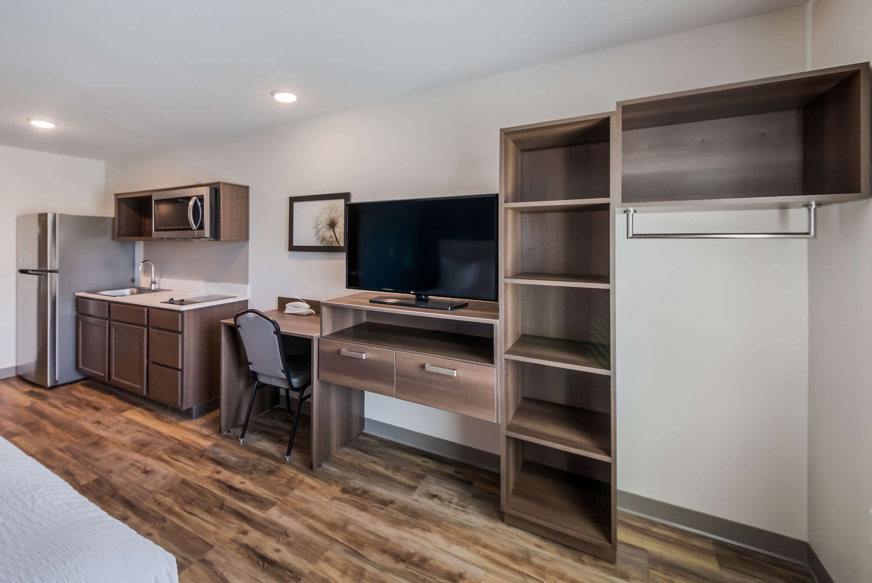 A modern bedroom with a bed, a desk with a chair, a television, and a kitchen area with a refrigerator and microwave.