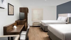 Ein Hotelzimmer mit Schreibtisch, zwei Betten, einem Fernseher und einem Fenster mit Jalousien.