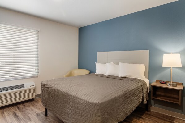 A hotel room with a bed, a chair, a nightstand with a lamp, and a window with blinds.